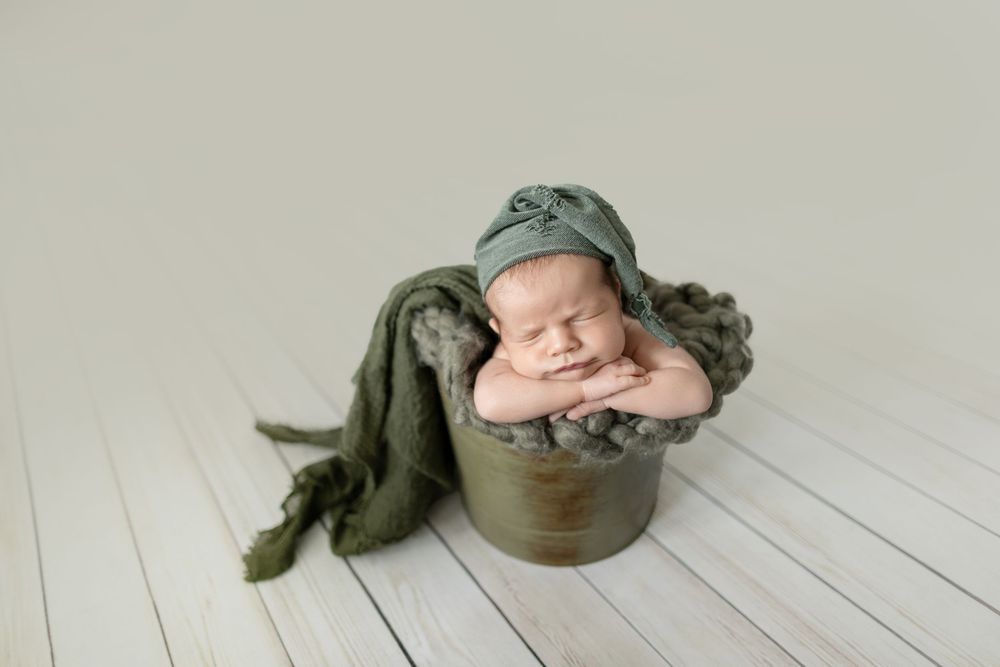 Newborn baby sleeping peacefully in a rustic bucket with green knit hat and wrap on white wood floor.