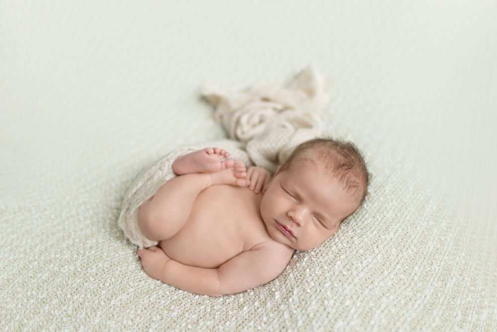 Peaceful newborn baby sleeping curled up on a soft cream blanket with a delicate floral wrap.