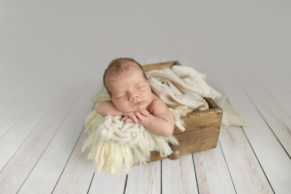 Sleeping newborn baby nestled in a rustic wooden crate with cream fluffy blankets on a white wood floor.