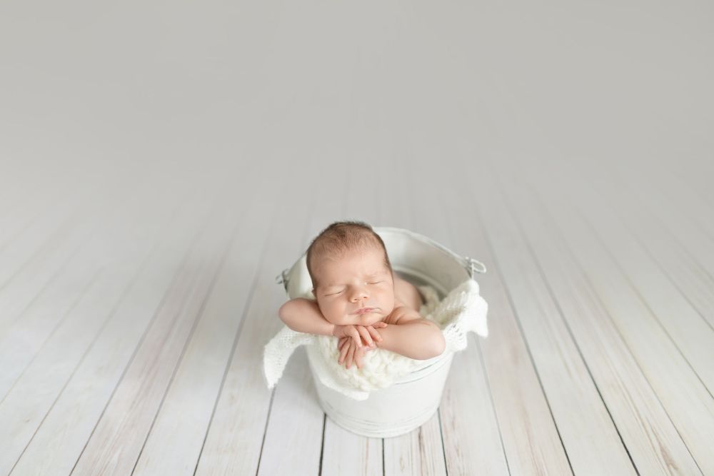Sleeping newborn baby posed in a white bucket with soft white fabric on a light wood floor.