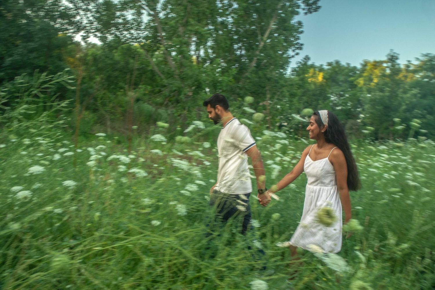 Couple in white outfits holding hands walking through lush green wildflower meadow on a sunny day.