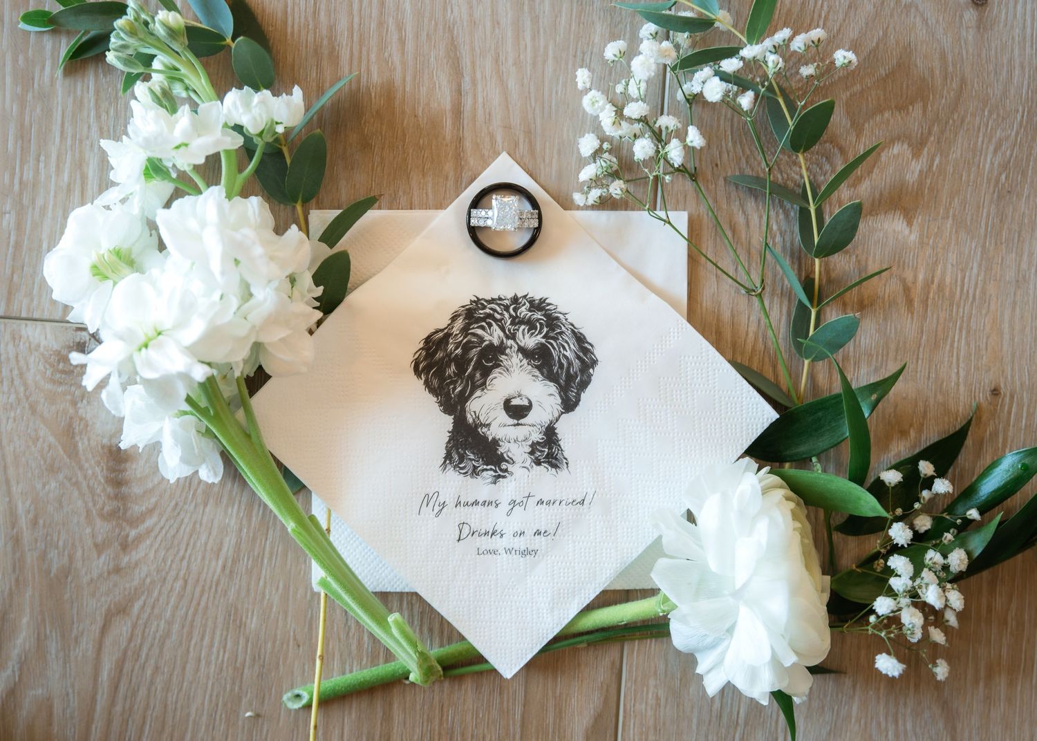 Wedding handkerchief printed with a sketched curly-haired dog and a message, surrounded by white flowers and greenery on a wooden table.