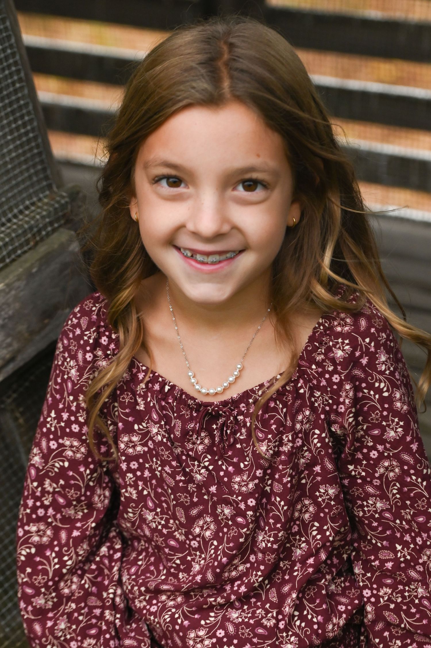 Smiling young girl with long wavy brown hair wearing a burgundy floral blouse and a beaded necklace, posing outdoors near rustic wood.
