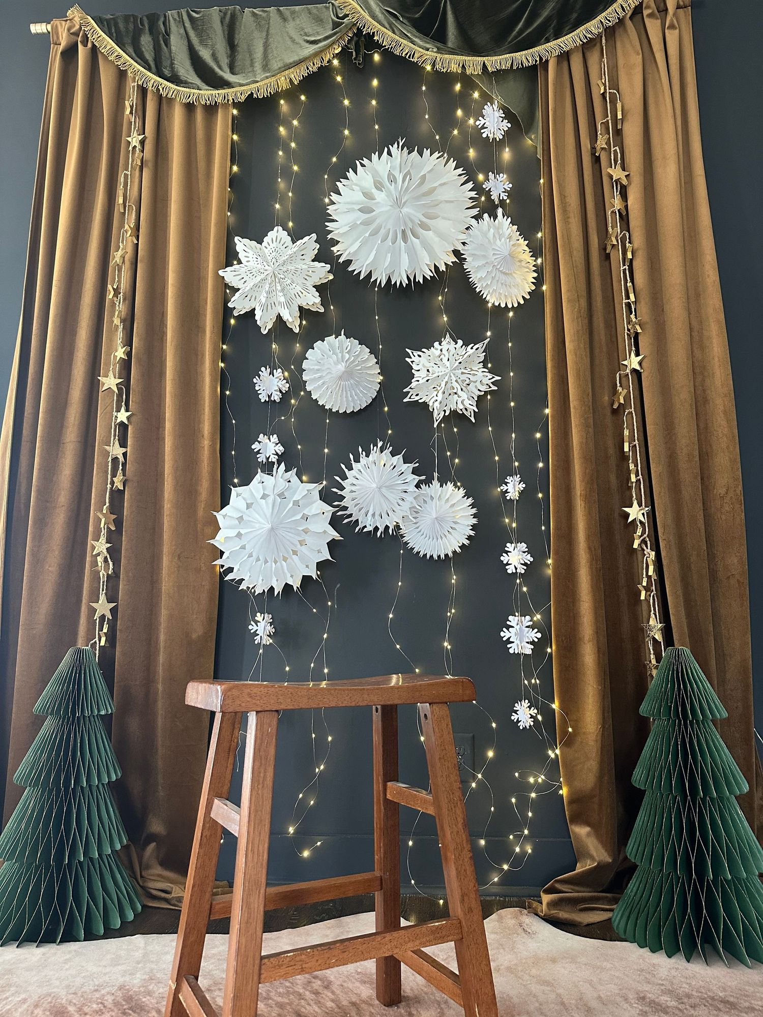 Festive Christmas backdrop with white paper snowflakes, fairy lights, gold star garlands, green tree decorations, and a wooden stool.
