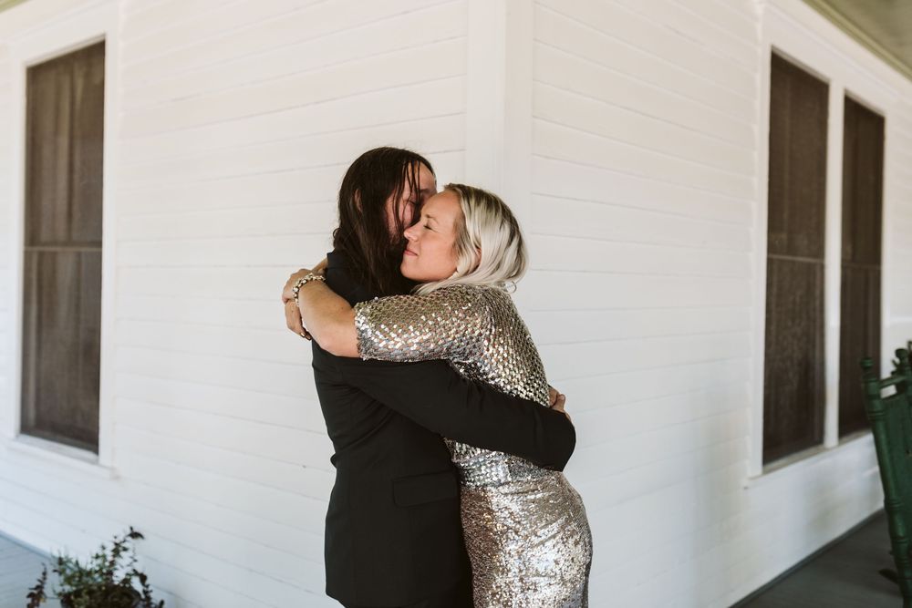 Two women sharing a warm hug outdoors, one in a black suit and one in a gold sequin dress.