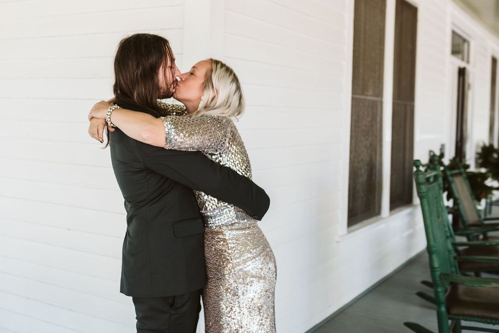 Couple embracing in a warm hug on a porch, woman wearing a sparkling sequin dress and man in a dark suit.