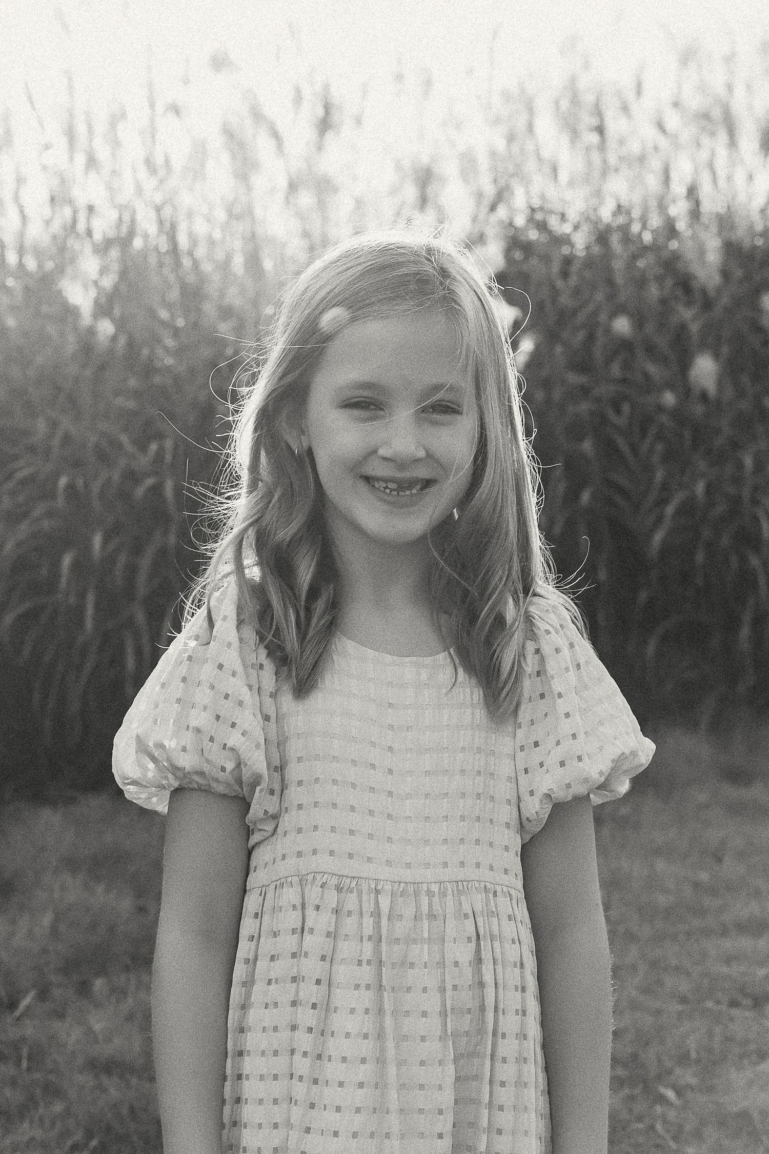 Black and white portrait of a smiling young girl in a dress outdoors with tall grass in the background.