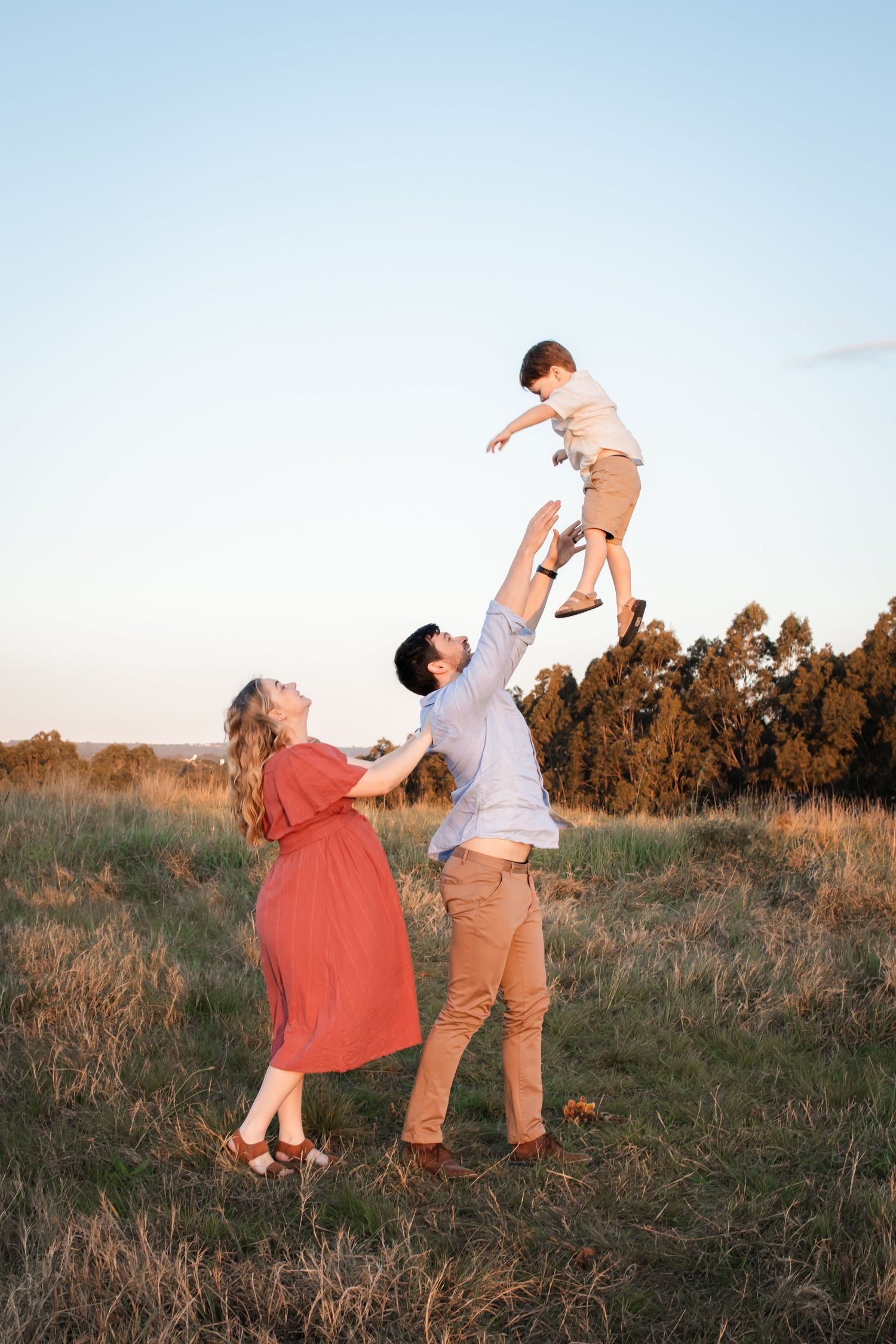 A joyful family outdoors at sunset, father tossing young boy in the air while mother watches in a red dress at Rouse Hill Regional Park.