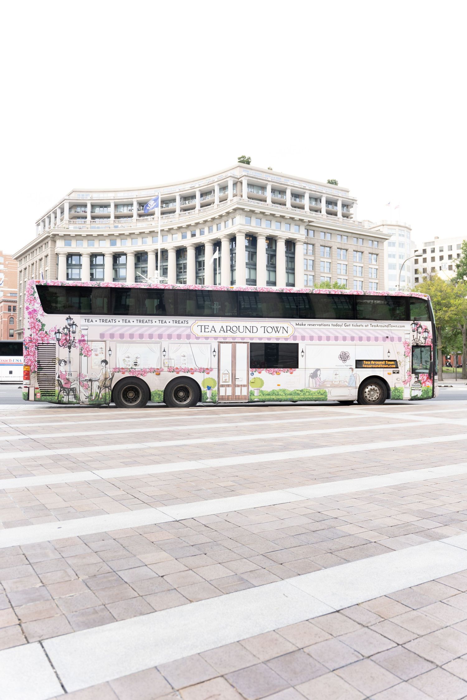 Pink Tea Around Town double-decker bus parked in urban plaza with historic building backdrop.