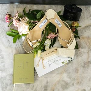 Gold Jimmy Choo wedding sandals with floral arrangement, monogrammed journal, and elegant bridal accessories on marble surface.