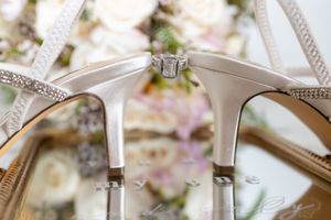 Elegant white satin bridal heels with rhinestone details displaying a diamond engagement ring, with soft floral bouquet in background.