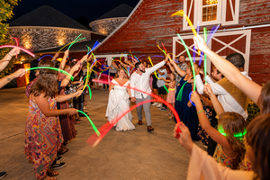 Bride and groom exit wedding reception through tunnel of colorful glowing neon sticks held by guests at night.