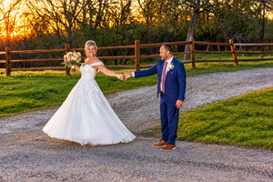 Bride and groom hold hands at sunset on a gravel path, with a wooden fence and golden trees in the background.