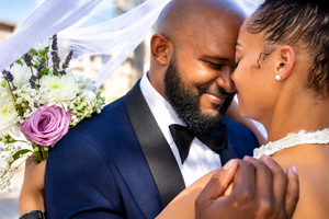 Bride and groom share an intimate moment under a veil, groom in navy tuxedo holding a purple floral bouquet.