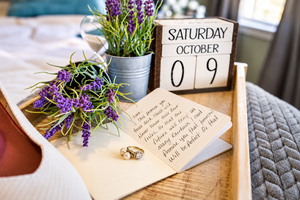 Wedding rings and handwritten vows on a wooden tray with lavender flowers and a calendar showing Saturday October 9.