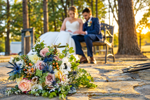 Wedding bouquet with roses and thistles in foreground, bride and groom sitting on bench at golden hour sunset.