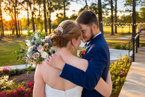Bride and groom share a romantic embrace at sunset, surrounded by pine trees and string lights at an outdoor wedding venue.