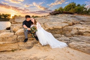 Bride and groom share a romantic kiss on rocky terrain at sunset, bride holding floral bouquet.