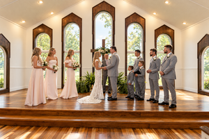 Wedding ceremony in elegant chapel with arched windows, bride and groom at altar with bridesmaids and groomsmen in pink and gray.