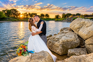 Bride and groom kissing by rocky lakeside at sunset with colorful bouquet, golden sky reflecting on water.