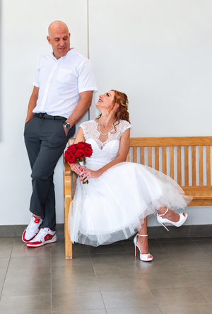 Bride in white dress holding red roses sits on wooden bench beside groom in white shirt and red sneakers.