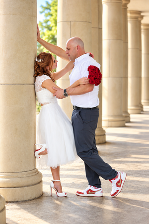 Bride and groom share a romantic moment between stone columns, holding red roses, wearing white outfits and red Nike sneakers.