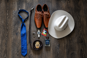 Flat lay of mens accessories including blue tie, brown oxford shoes, white cowboy hat, watch, cufflinks, belt, and cologne.