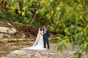 Bride and groom embrace by a rocky creek surrounded by lush green trees, bride's long veil flowing behind her.
