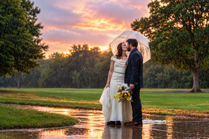 Bride and groom share a romantic moment under a clear umbrella in the rain at sunset on their wedding day.