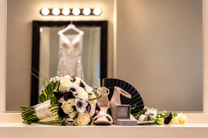 Bridal flat lay featuring a white bouquet, heels, perfume, and accessories on a vanity with a wedding dress reflected in the mirror.