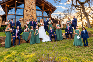 Wedding party poses outside stone venue, bridesmaids in green dresses and groomsmen in navy suits with pink ties.