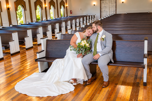 Bride and groom share a tender moment sitting on church pews, bride in white gown holding bouquet.