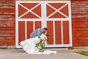 Bride and groom sharing a romantic dip kiss in front of a rustic red barn with white cross doors.