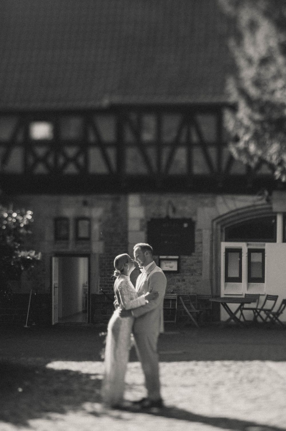 Black and white wedding photo of couple embracing and kissing in front of a rustic half-timbered building.