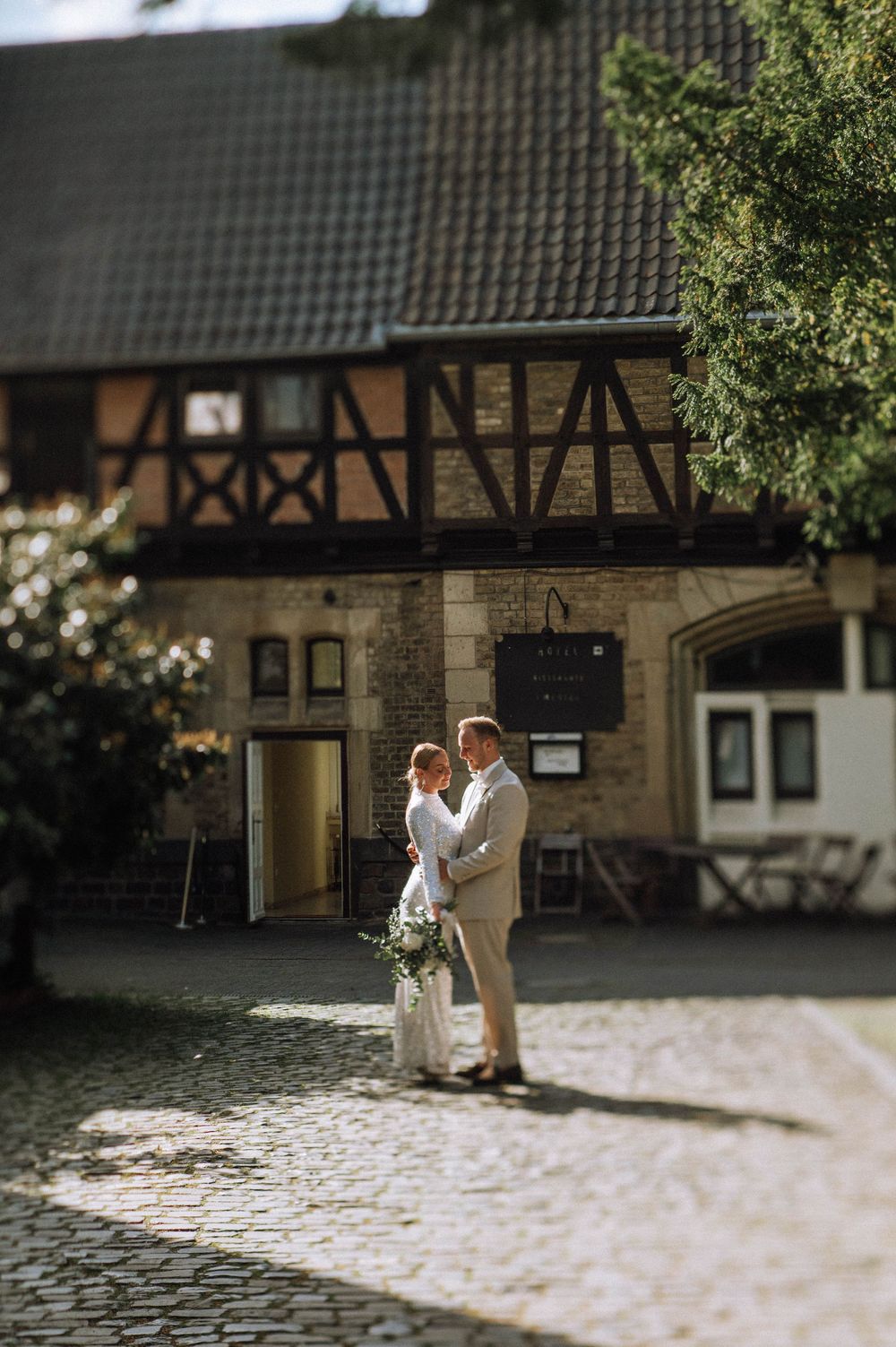 Bride and groom embrace on cobblestone courtyard before a charming half-timbered building on their wedding day.