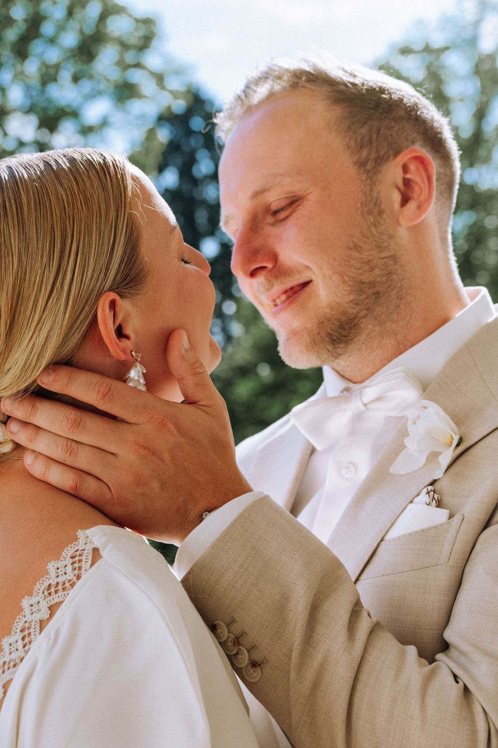A groom in a tan suit tenderly holds his bride's face as they share an intimate moment outdoors on their wedding day.