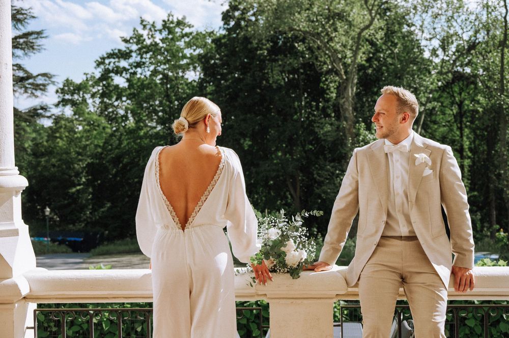 Bride in backless white gown and groom in beige suit share a romantic moment on a sunlit balcony.