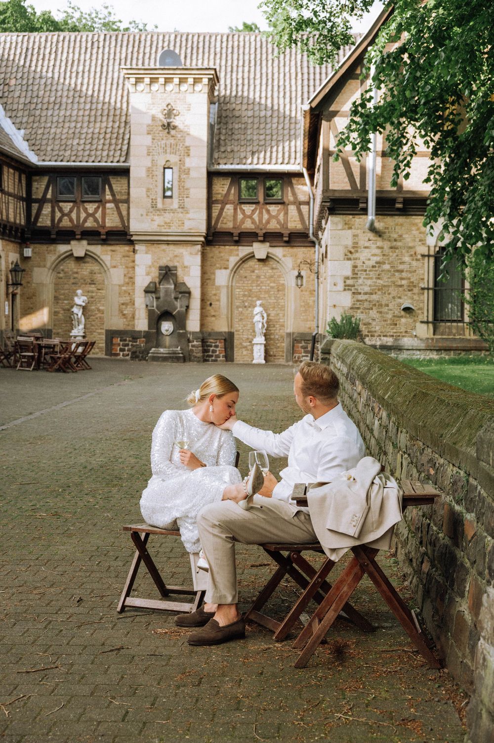 Bride and groom in white attire sit on wooden chairs by a stone wall at a historic European venue.
