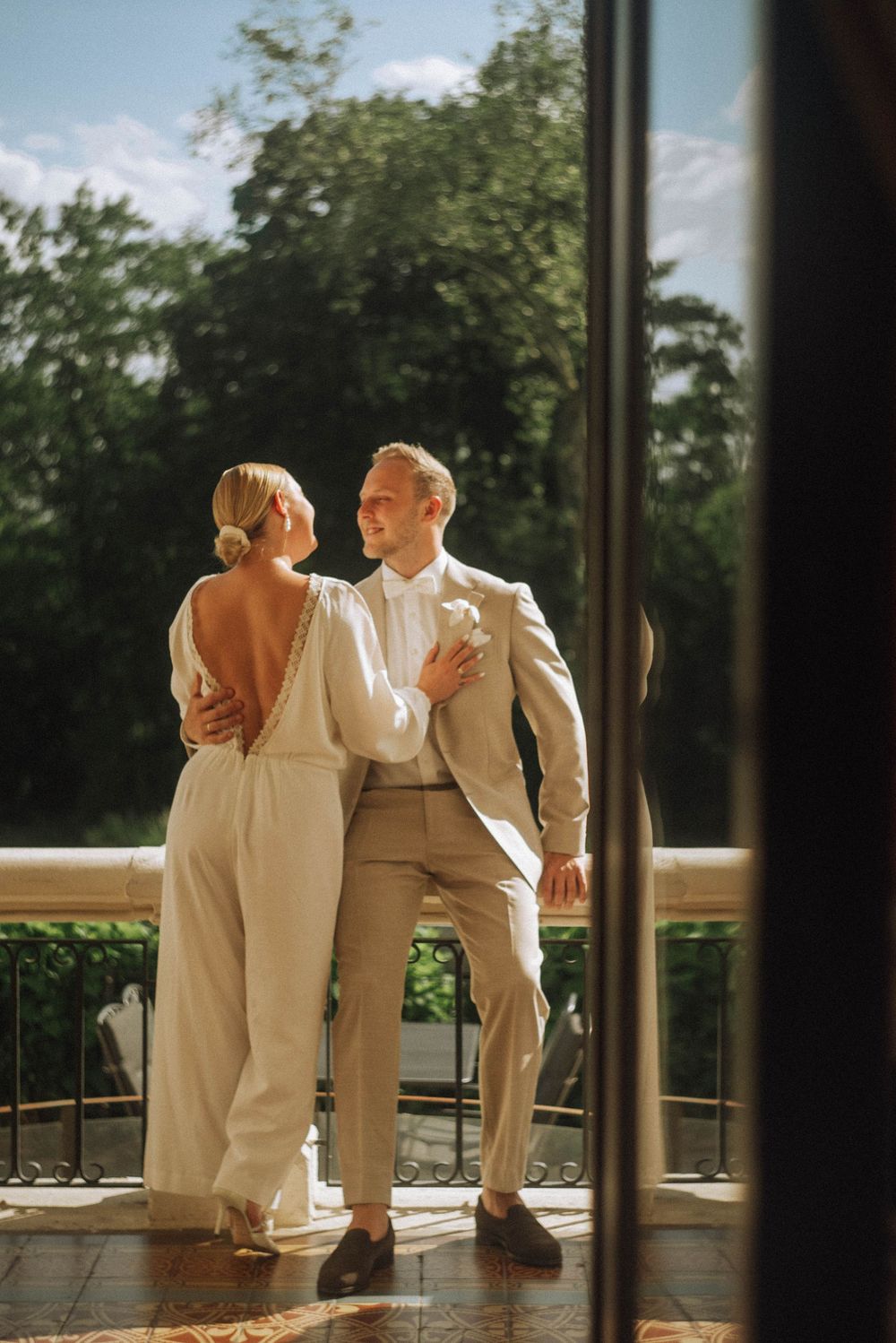 Bride and groom embrace on a sunlit balcony, she in a backless white jumpsuit, he in a beige suit, with lush greenery behind.