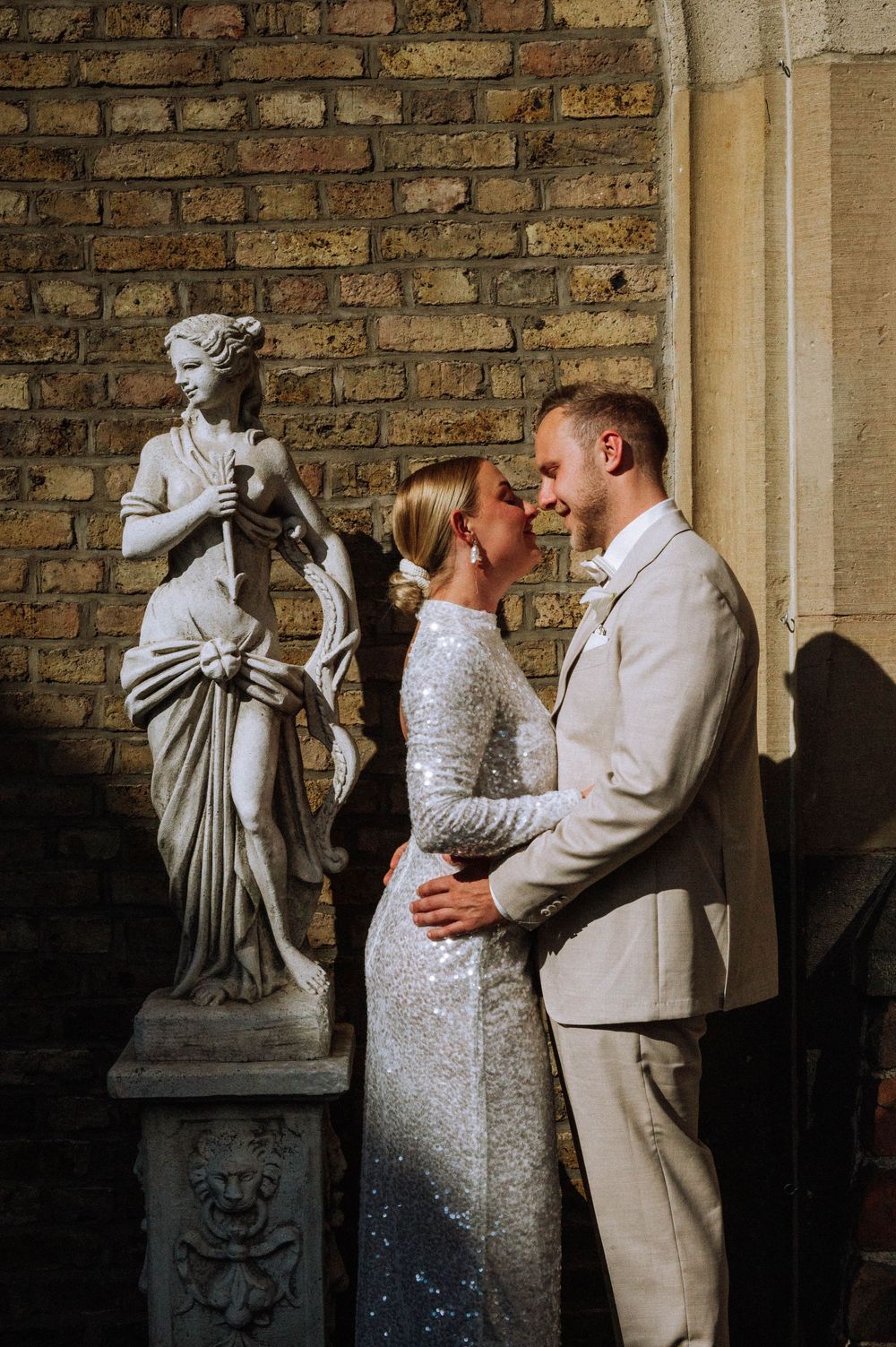 A bride in a sequin gown and groom in beige suit share a romantic moment beside a classical statue against a brick wall.