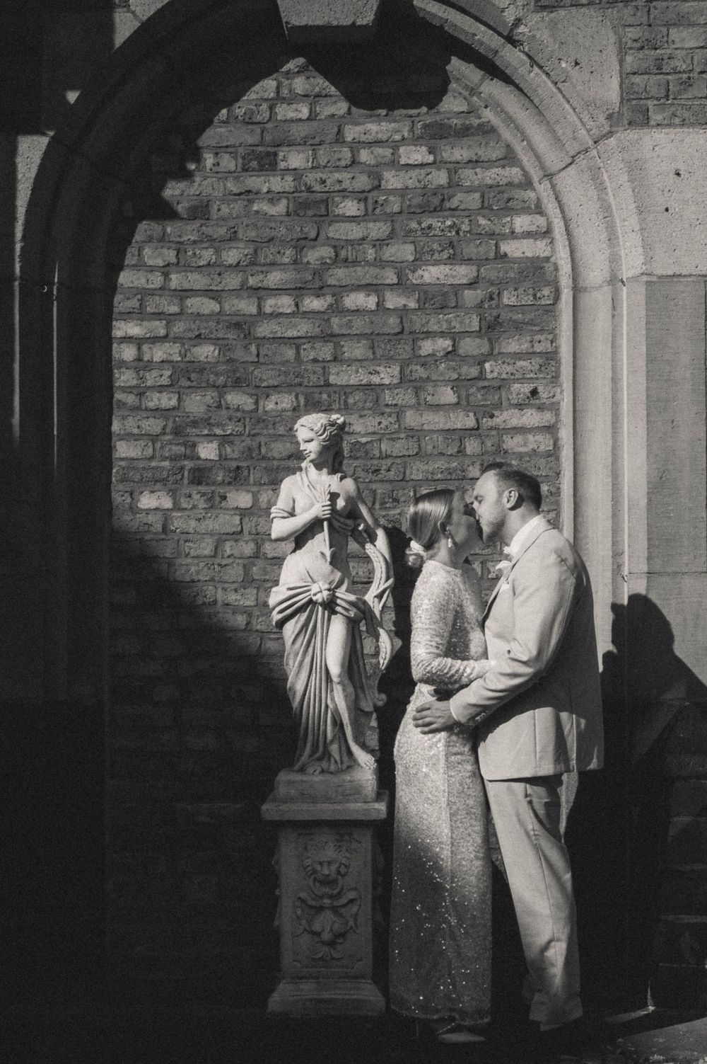 Black and white photo of a couple sharing a romantic moment beside a classical statue under a brick archway.