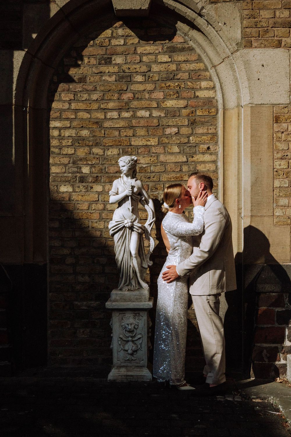 Bride and groom sharing a romantic kiss beside a classical statue under a stone arch with brick wall backdrop at Schlosshotel Kommende Rame