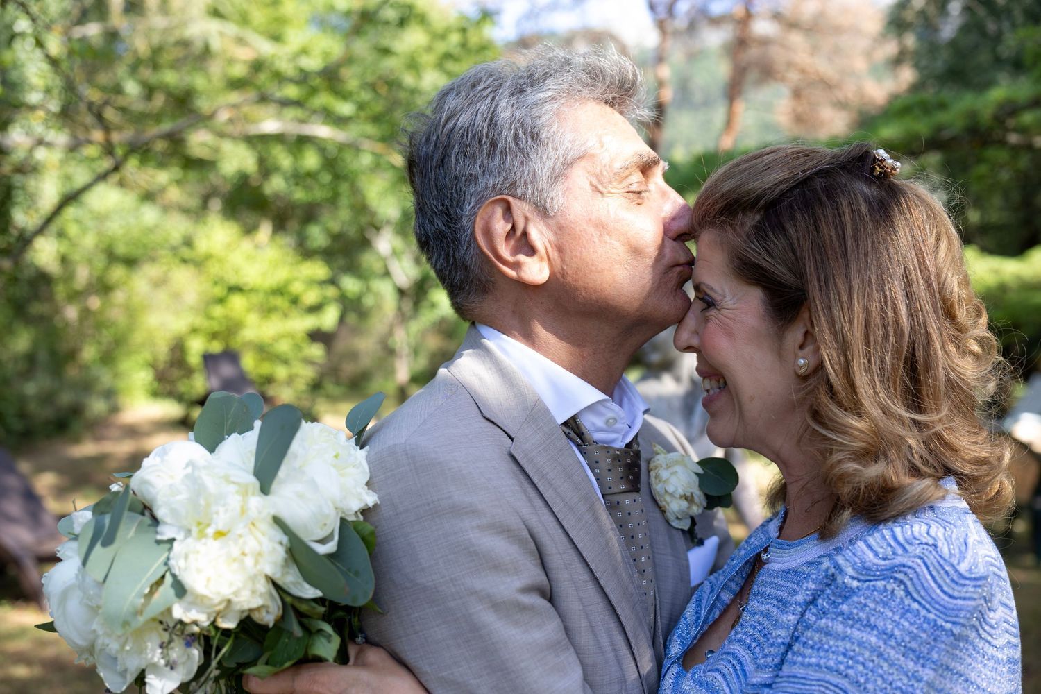 Le marié embrasse le front de la mariée, bouquet à la main, devant les arbres