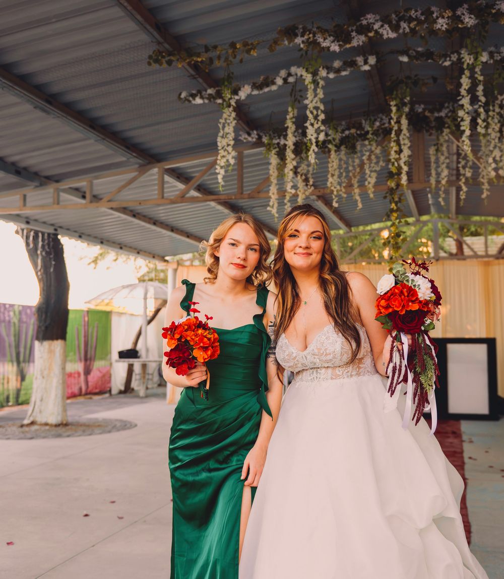 Bride in white gown and bridesmaid in emerald green dress holding red bouquets at an outdoor wedding venue.