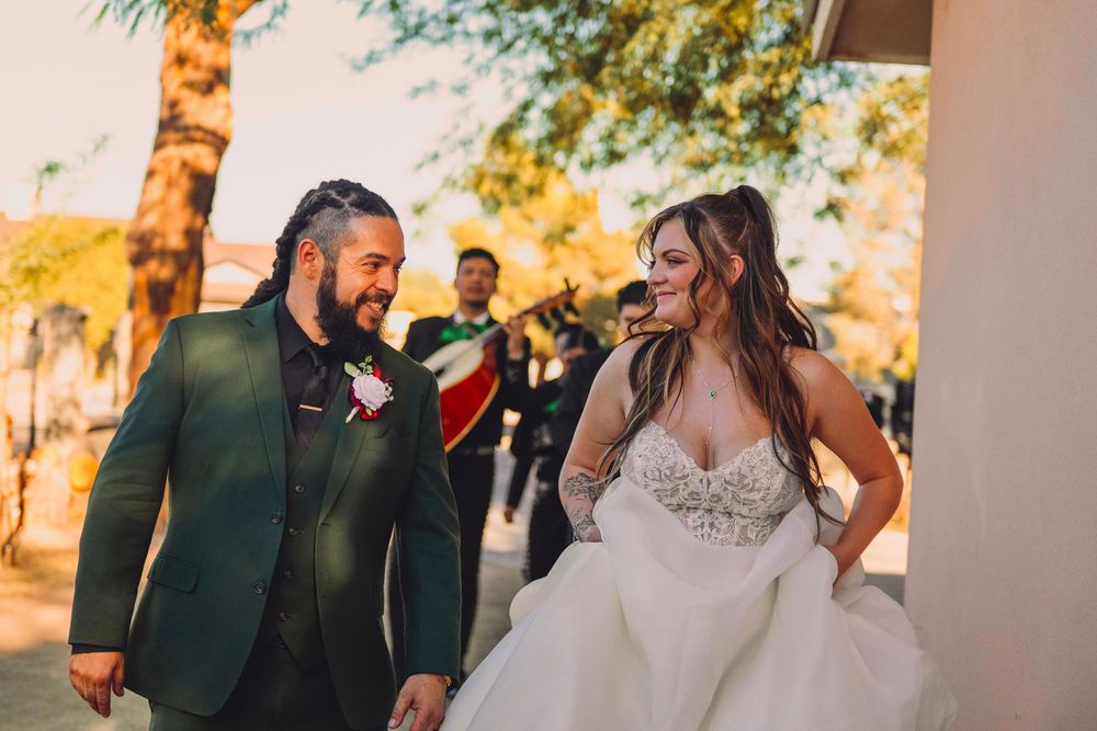 Bride and groom laughing together outdoors at wedding with mariachi band in background.