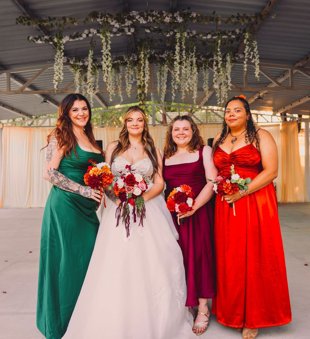 Bride in white gown with three bridesmaids in green, burgundy, and red dresses holding bouquets at wedding venue.