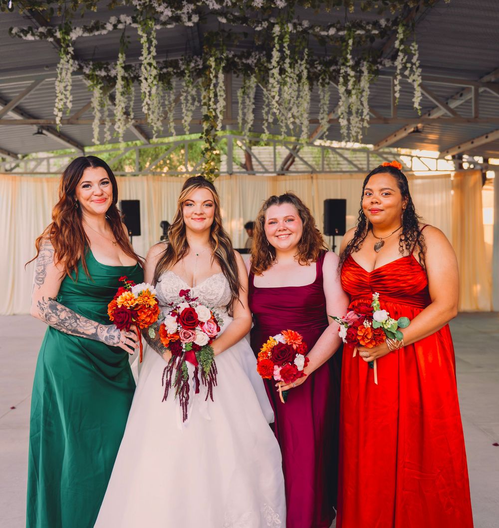 Bride poses with three bridesmaids in green, purple, and red gowns holding bouquets at wedding reception.