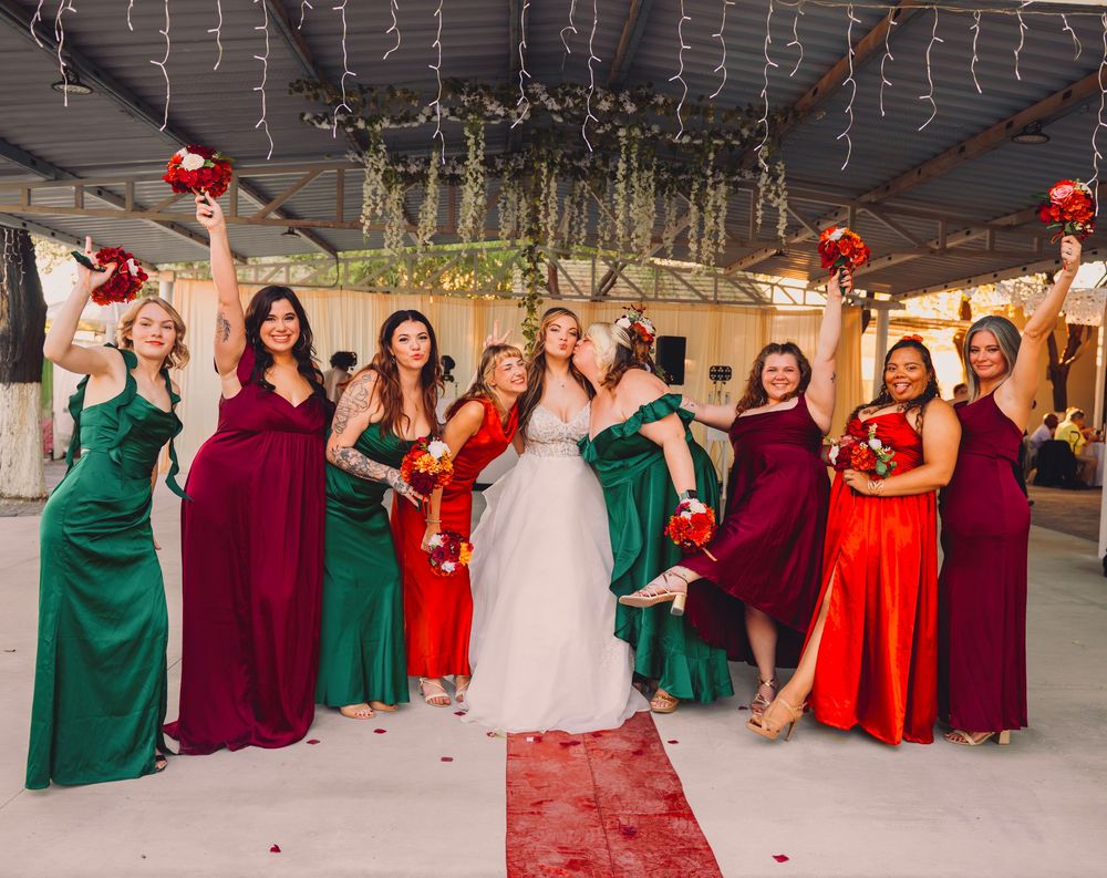 Bride poses with bridesmaids in green, burgundy, and red gowns holding bouquets at elegant wedding venue.