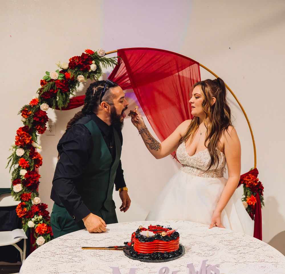 Bride feeds groom cake at wedding ceremony under red floral arch with red velvet cake on table.