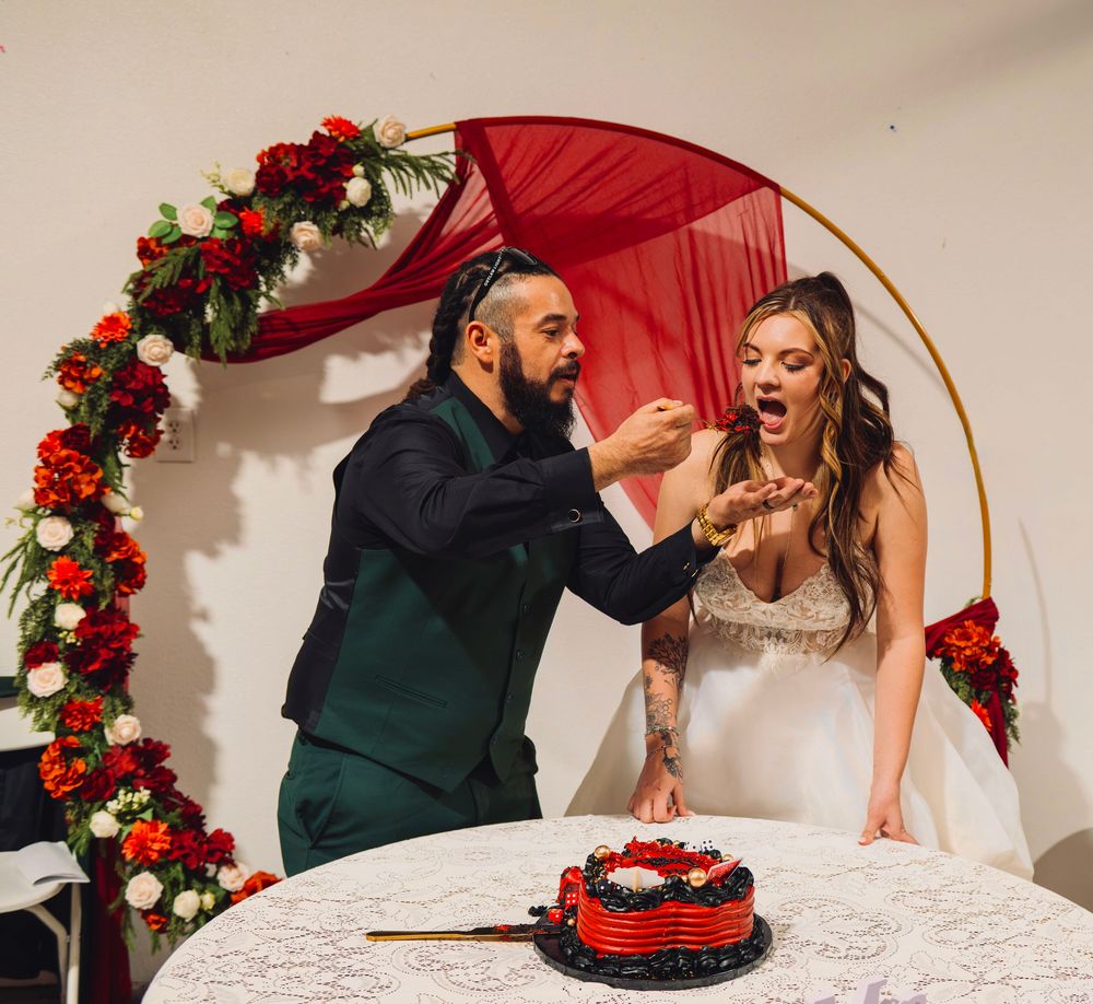 Bride and groom cutting a red wedding cake under a floral arch with red roses and a red fabric backdrop.